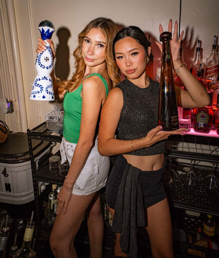 Two professional female bartenders posing back-to-back with premium tequila bottles ready to serve